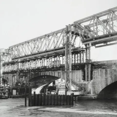 The London Archives, City of London A black and white image showing the a metal frame in place of the bridge, during construction