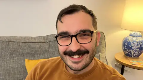 Guto has short dark hair, thick eyebrows, black rimmed glasses and a thick moustache, as well as some shorter black beard hair. He is smiling at the camera and is wearing a yellow sweatshirt