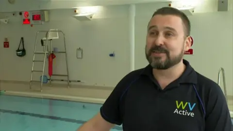 A man stands next to a pool with a lifeguard chair in the background. He has short brown hair and is wearing a black polo shirt with a WV Active logo on it.