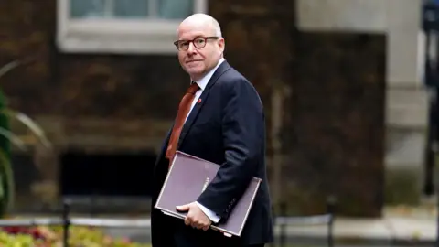 PA Media Attorney General Richard Hermer arrives at Downing Street. He is wearing a dark suit and burnt orange tie with a small poppy on his left lapel. He is carrying a dark red file in his hand.