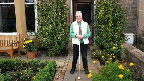 Lorna Pratt Lorna Pratt standing in a well-kept garden that has bushes and yellow flowers and a creeping plant that is growing up both sides of the front of the house she is outside. She is standing on a path that leads to the front door. She is holding her white cane and is wearing a white body warmer, black trousers and glasses.