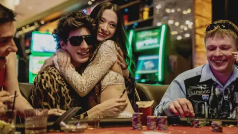 Neon Mikey Madison and Mark Eidelstein in Anora. Mark Eidelstein's character is sitting down at a poker table and is wearing glasses and leopard skin jacket. Mikey Madison is wearing a cream coloured dress and sitting on his lap. They are both smiling. 