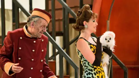 KK Dundas A scene from Scottish Opera's 2014 version of Don Pasquale,  as the character Norina - in a multi-coloured dress, holds Bridget the dog - dressed as a cat, while a porter looks on