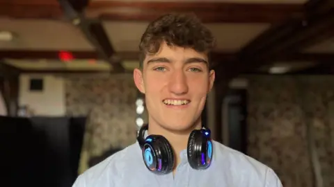 Matthew Harris, a student at the RAU waring a blue shirt and big black headphones. Behind him is dark as he is at a silent disco.