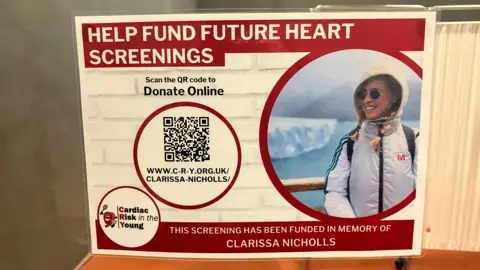 Robbie Kalus/BBC A red and cream poster, containing a photograph of Clarissa Nicholls, inviting people to "help fund future heart screenings"