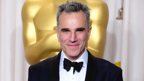 PA Media Daniel Day-Lewis, dressed in a dinner suit, holding an Academy Award for Best Actor while standing in front of a large golden Oscar statuette.