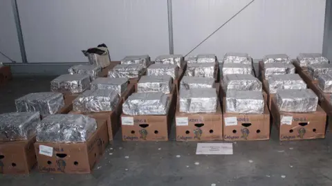 Crown Office Dozens of cardboard boxes lined up on the floor of a warehouse. There are silver packages on top of every box.