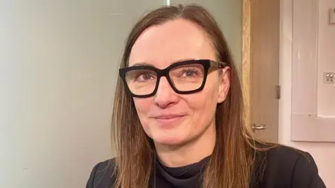 Sineidin Corrins in a room with plain beige walls and a door behind her. She is wearing dark-rimmed glasses and a black top