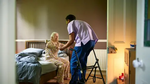 Getty Images A care home nurse in a light purple tunic and blue trousers helping an elderly woman in brown pyjamas out of bed. There is a walker positioned nearby and the door is open with the light from a lamp reflected on the door, giving a warm glow.