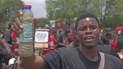 A protester wearing a black T-shirt holds up a bottle of dirty water. Other people can be seen behind him