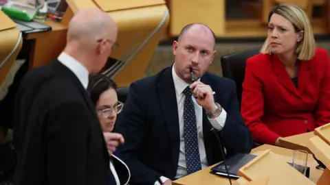 PA Media Neil Gray sitting inside the Scottish Parliament chamber with a pen in his mouth and a folder on the desk. He is wearing a dark suit and tie and is listening to John Swinney who has his back to the camera. Jenny Gilruth is sitting next to Gray wearing a bright red jacket and Kate Forbes sits between Swinney and Gray.