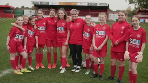 Shaun Whitmore/BBC Female coach surrounded by girls wearing red tops and shorts and football boots.