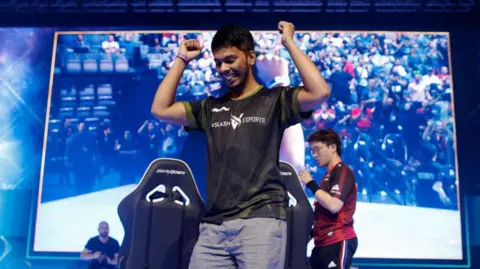 Getty Images A young man stands in front of a giant screen, smiling with his arms held up in a victory pose. Two gaming chairs, facing the screen, are just behind him. 