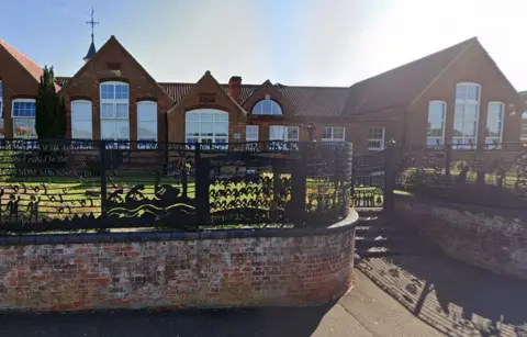 Google Black fences surround Fakenham Juniors, with grass in the foreground of the red brick building.