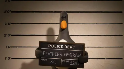 Feathers McGraw, a stop-motion animated penguin, is seen holding up a police department sign while having a mugshot taken