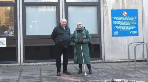 Iain Young Iain Young is standing outside the hospital with a friend. He is in a black jacket and black trousers and he is resting his hands behind his back. His female friend is in a long green winter coat and has a white cane.