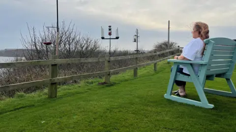 Kevin Shoesmith/BBC  Angie Moore sits in a pale green, wooden chair on grass overlooking the sea. On the far left of the picture, is a headland.