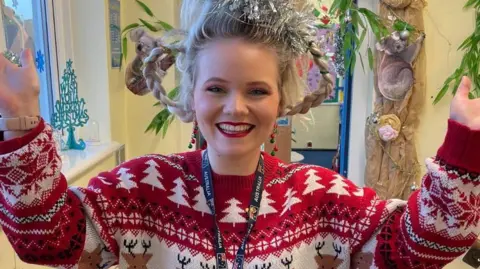 Hannah Gidman Hannah Gidman smiles at the camera with her hands up. She is dressed in a Christmas jumper with her hair in plaits and wrapped in tinsel. 