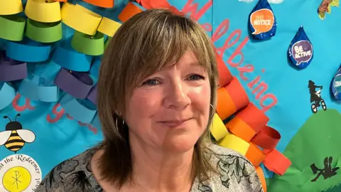 Andrea Adams, who is attending the classes to help her grandson, stands with a smile, wearing a grey flowery dress and hoop earrings. She has short brown hair and is standing in front of a colourful wall pattern.