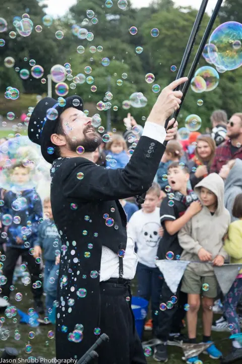 Jason Shrubb Man in a top hat juggles bubbles with a wand as a crowd watches on