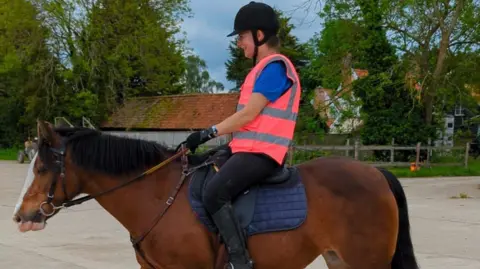 Amy Anderson Amy in a high visibility vest and sat on a brown horse