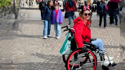 Amy Anderson Amy in a red coat and wearing a Minnie Mouse head piece while sat in her wheelchair at Disney Land