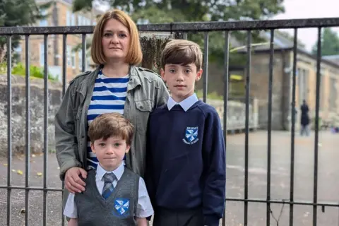 Julie, wearing a stripy top and a green jacket, stands with her arms around her songs in their primary school uniform at the school gate.