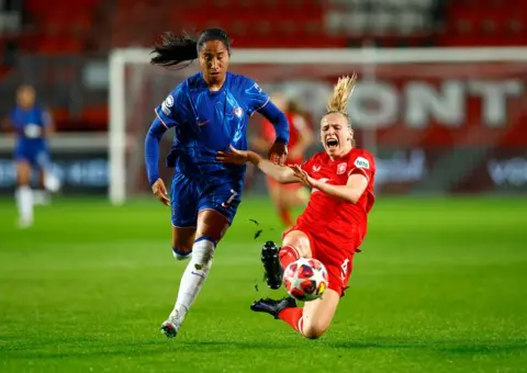 Piroschka Van De Wouw/Reuters A woman with dark long hair, wearing a blue football kit with white socks runs towards a ball and a woman with fair hair wearing a red football kit lunges with her right foot towards the ball. The woman in red is shouting as she falls towards the grass pitch. 