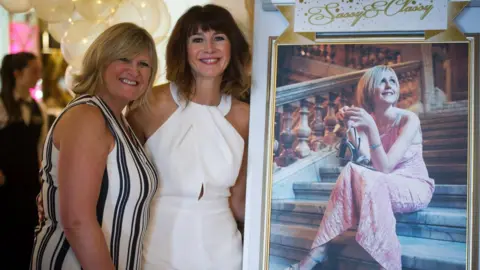 Angela MacVicar Smiling Angela MacVicar and Lindsay MacCallum wearing party dresses posing next to a photograph of Johanna MacVicar with helium-filled balloons behind them