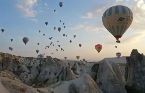 Behcet Alkan/Anadolu via Getty Images Hot air balloons fly over Cappadocia, a UNESCO World Heritage Site.  on October 14, 2024.  Cone-shaped rock formations are in the foreground.  Over twenty balloons of various colour, rise up at various distances into a pale blue sky.  Faint clouds are on the right hand side.