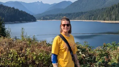 Amy Anderson Amy wearing a yellow t-shirt and stood by a lake with mountains in the background