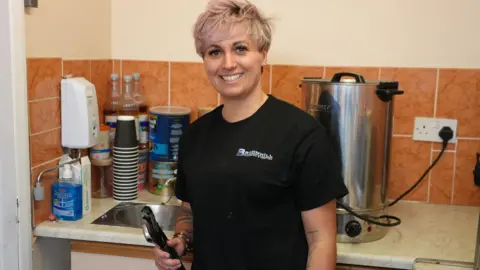 Shaun Whitmore/BBC Woman is smiling and has pink hair. She is in a kitchen with a tea urn and paper cups and a sink behind her. She is wearing a black T-shirt and holds a large spanner.