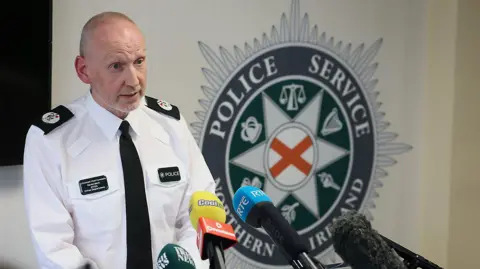 Pacemaker A police officer, wearing a phone shirt and black tie, standing in front of media microphones. There is a PSNI logo on the wall behind him. 