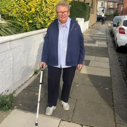 Hazel Kelly Hazel Kelly is walking along a street with the aid of a white walking stick. She is wearing glasses and a blue jacket and pale blue shirt and black trousers.