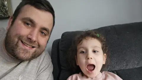 Columba Mailey Columba and Maisie together on a grey sofa, Columba is smiling and wearing a grey sweatshirt and Maisie has her mouth open wide while wearing a pink sweater