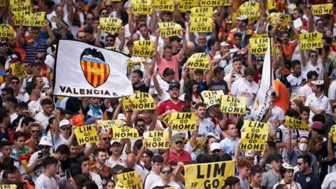 Getty Images Hundreds of people, several of whom are holding 'LIM GO HOME' signs, protest before a game in Valencia