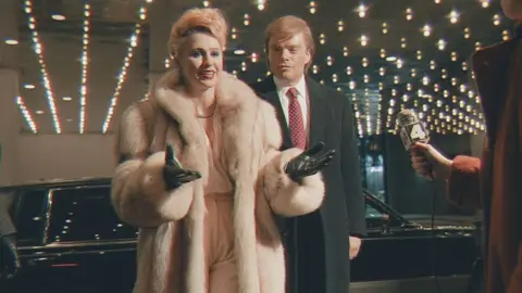 Studio Canal Maria Bakalova and Sebastian Stan as Ivana and Donald Trump in The Apprentice - Bakalova is wearing a fur coat and black leather gloves, Stan has a red tie, white shirt and black overcoat. They are being interviewed by a reporter.