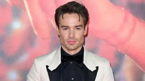 Getty Images Liam Payne has brown hair and is wearing a white blazer and a black shirt. He is standing in front of a red background with a slight smile on his face. 