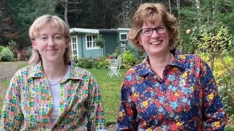 Jenny Gow Eilidh and her mum Jenny. Eilidh has blonde shoulder length hair and she is wearing patterned pink, green, yellow and purple dungarees. She is stood in a garden with a summer house in the background. Jenny is smiling beside her wearing blue, yellow and red floral patterned dungarees. She is wearing glasses and has brown hair.