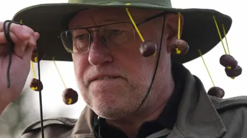 PA Media A man with glasses wearing a wide brimmed hat with conkers dangling from it