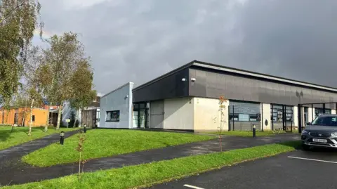 Exterior view of Bunscoil an tSléibhe Dhuibh in west Belfast.   It is a modern building surrounded by some trees, a lawn and paved walkways.  A car is parked in its carpark.