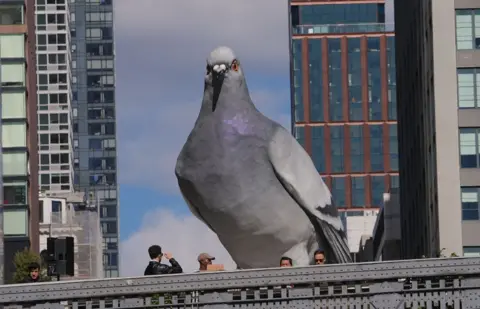 Selcuk Acar/Anadolu via Getty Images A large sculpture of a pigeon in the foreground with only the heads of people passing by visible below. Tall buildings are in the background and the sky is blue.