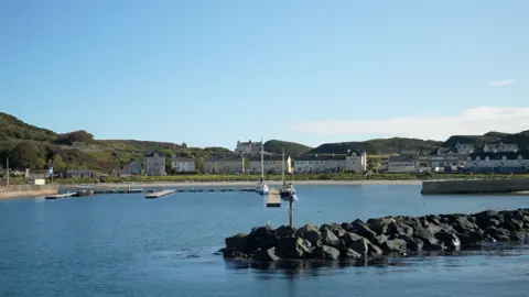 A view of Church Bay on Rathlin Island on a sunny day where a cluster of buildings line the shore around a harbour.  Two small boats are docked in the bay.