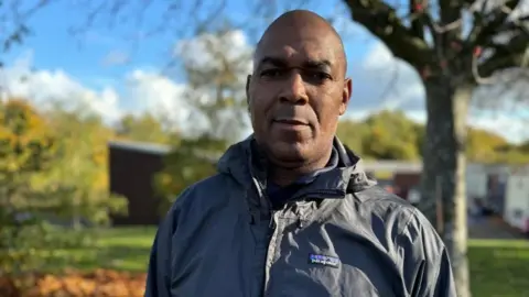 Jonny Humphries/BBC Rodney Tucker, wearing a grey Patagonia raincoat, looks into the camera with a serious expression against a backdrop of a grassy area and a tree