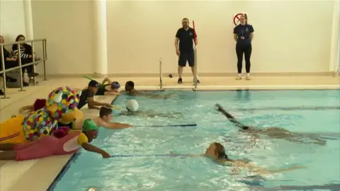 Four children lie down on the side of the pool, and are holding out long sticks to children in the water, who are reaching to them. There are two instructors watching on from behind the pool, and a large bull mascot is next to the children.