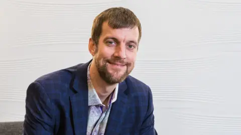 Jasco Headshot of Jason Trice smiling at the camera. He has brown hair and a brown beard and wears a shirt and jacket.