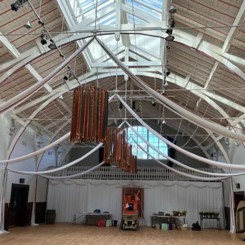 The hall has decorations hanging from the roof. There are tables and equipment at the back, along with the balcony and large window.