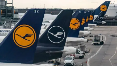 Getty Images The tail fins of eight Lufthansa aircraft parked at gates at Frankfurt airport.