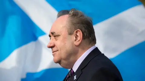 PA Media Alex Salmond is side-on to the camera with a Saltire flag behind him