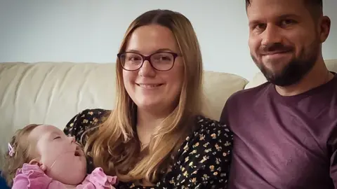 BBC Baby Robyn in a pink jumper. She has long blond hair, her eyes are closed and she has a breathing tube in her nose. Mum also has light hair a printed blouse. She is hold Robyn and smiling. Dad Daniel is in a T-shirt and is snuggled in to his wife on their couch